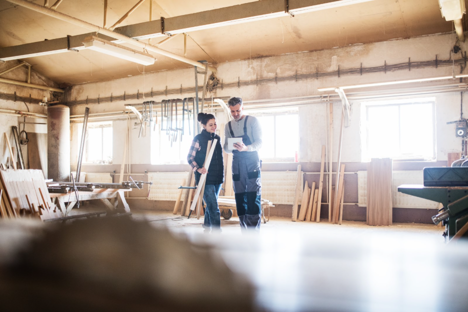 Contractors reviewing plans on a tablet inside an unfinished commercial workshop with exposed utilities, construction materials, and equipment.